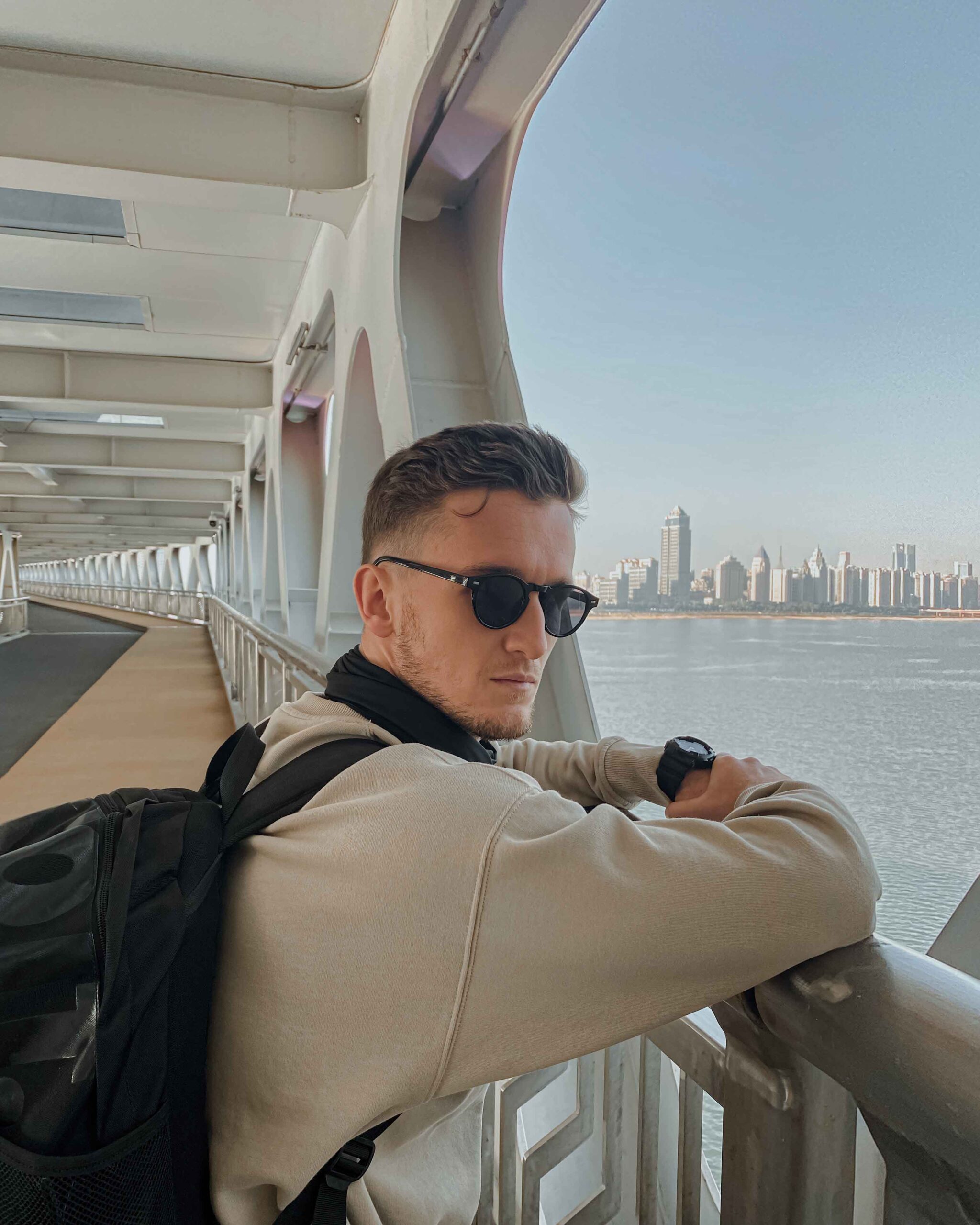 Man wearing sunglasses and a backpack standing on a bridge overlooking a city skyline by the river